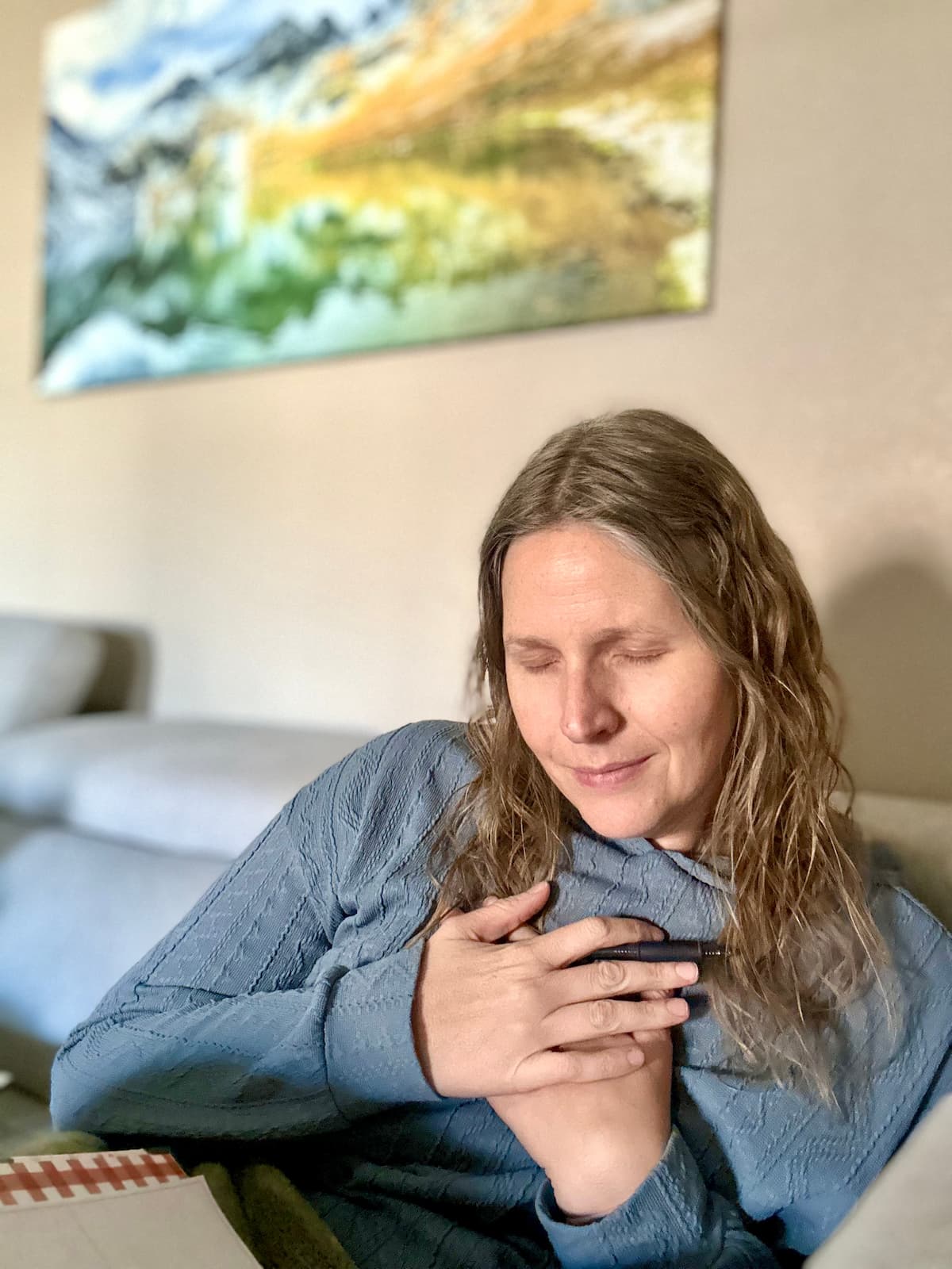 Woman sitting on her couch with her eyes closed hands gently folded over her heart, and eyes closed with a slight smile on her face as she prays